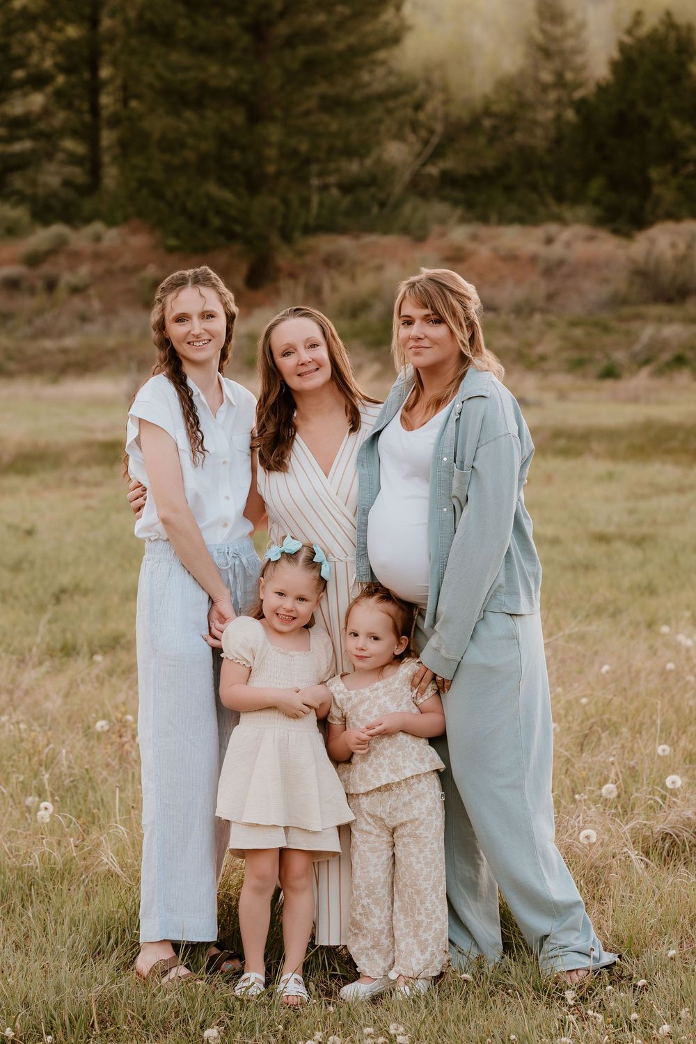 Family portrait of three women and two young girls posing together in a sunny meadow, one woman visibly pregnant.