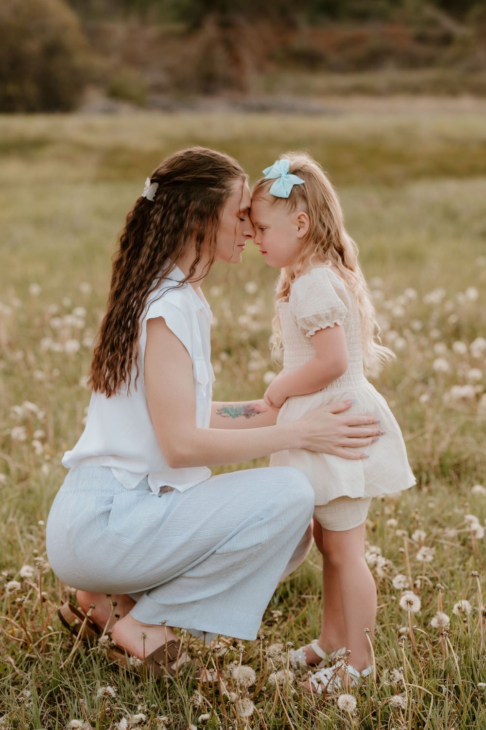 Mother and daughter touching foreheads in a dandelion field, sharing a tender moment outdoors.