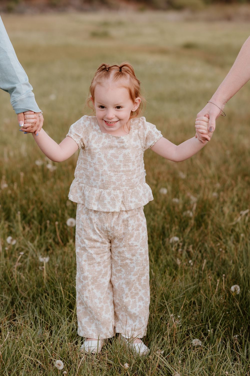 A smiling toddler girl in a floral outfit holds hands with two adults in a grassy field.