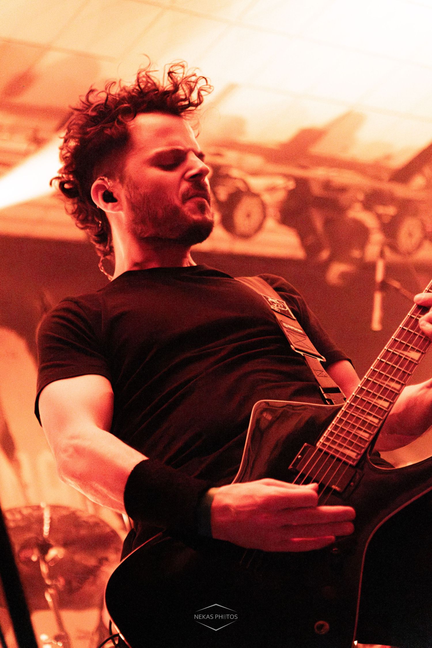 Male guitarist with curly hair plays electric guitar on stage under warm orange lighting during a live rock concert performance.
