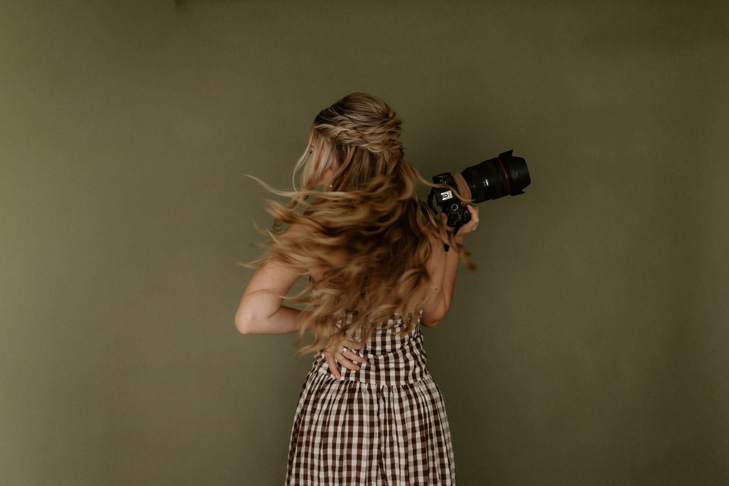 Young woman in plaid dress smiling, holding black hat, posing against olive green background.