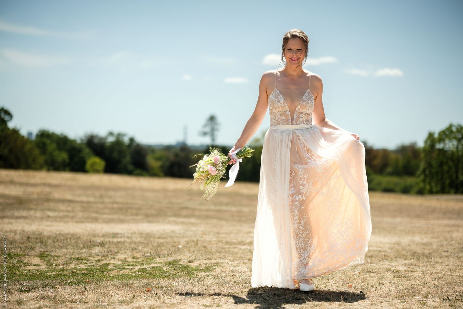 Bride in lace wedding gown holding bouquet stands in sunny field with blue sky backdrop.