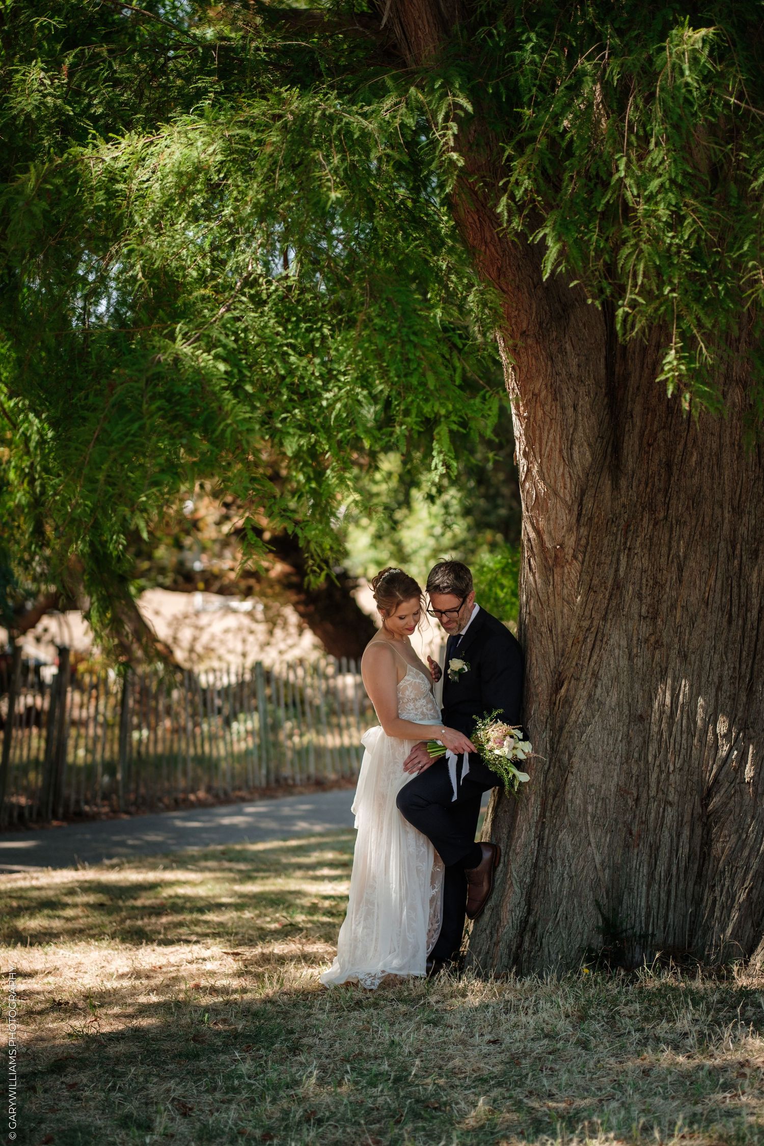 Bride and groom embrace under a large tree in an outdoor wedding portrait setting.