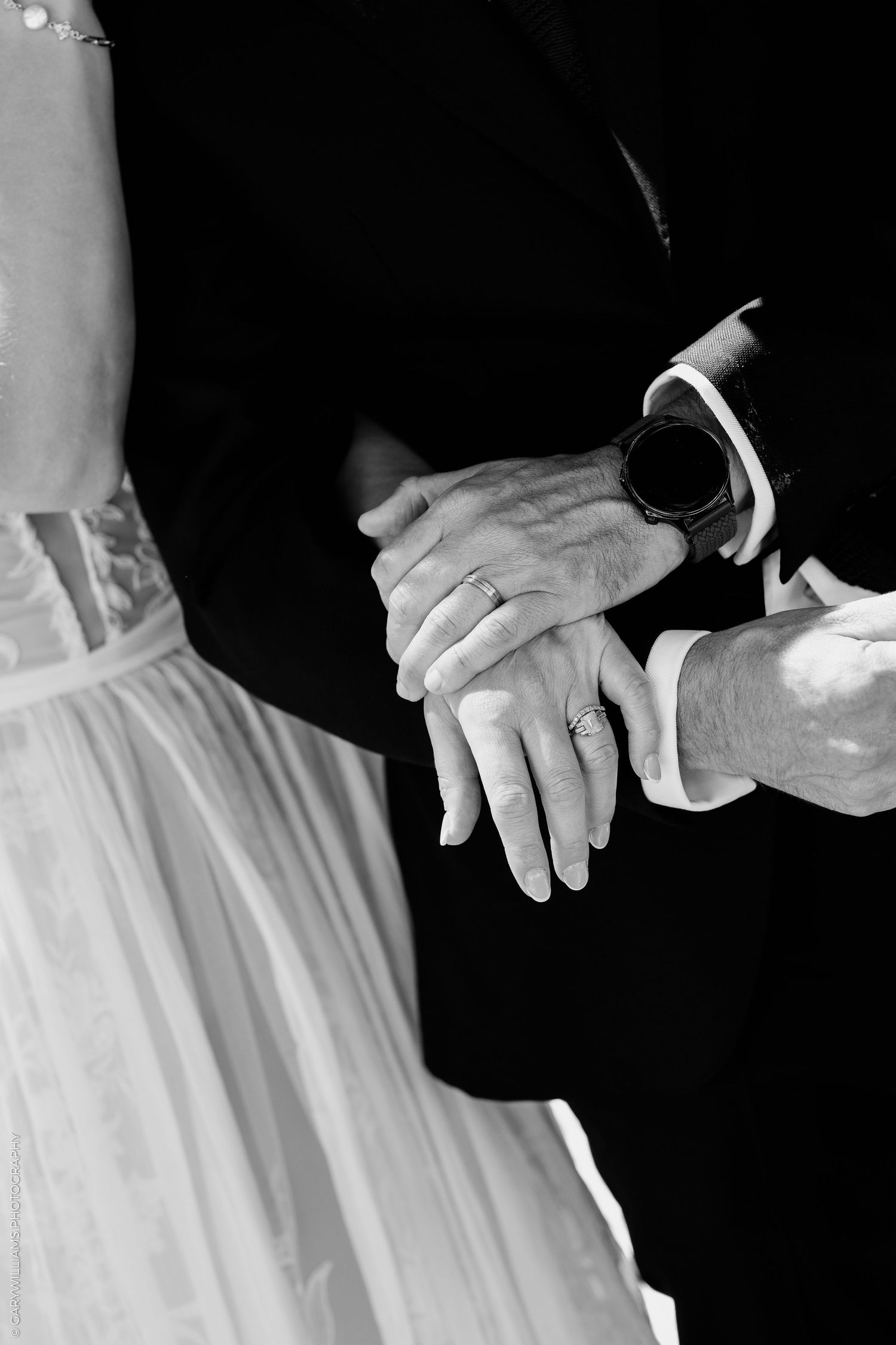 Black and white photo of bride and groom exchanging rings, close-up of hands with wedding bands and smartwatch.