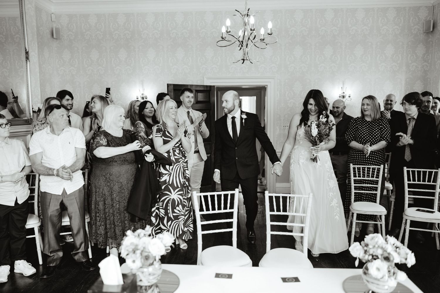 Black and white photo of a bride and groom making their grand entrance at a wedding reception with guests cheering.