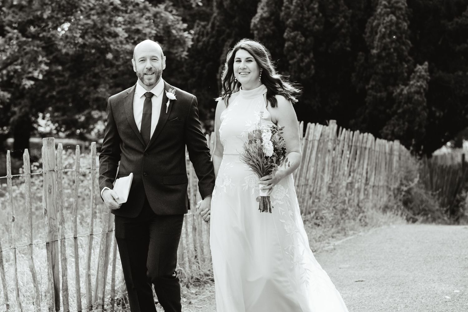 Black and white photo of a bride and groom holding hands walking along a rustic fence outdoors on their wedding day.