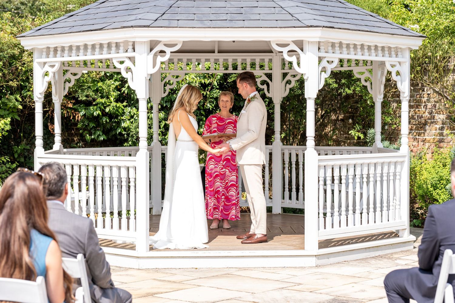 Couple exchanging vows in a white gazebo during an outdoor wedding ceremony with guests seated on a sunny day.