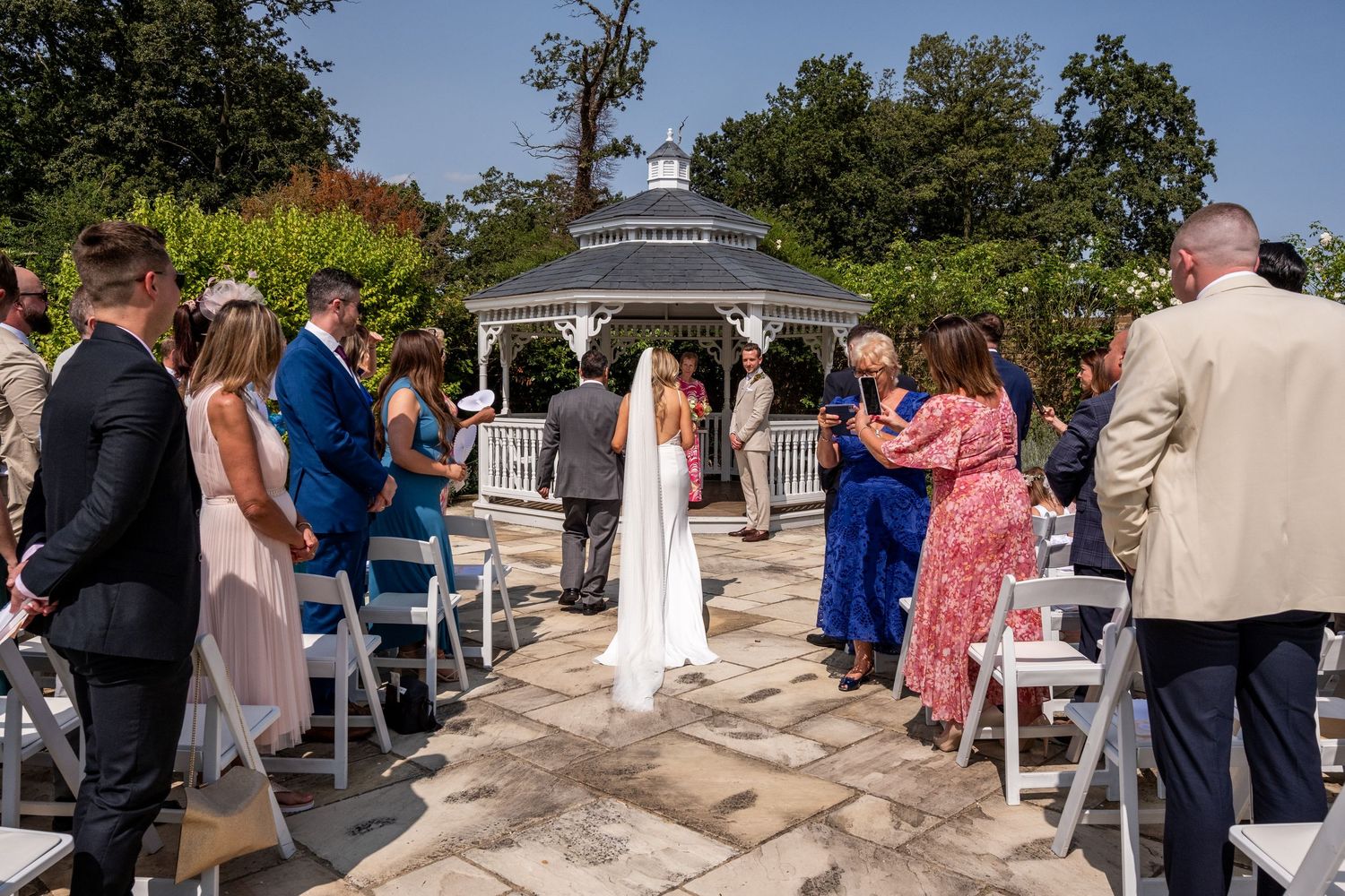 Outdoor wedding ceremony at a gazebo with guests seated in white chairs on a sunny day.