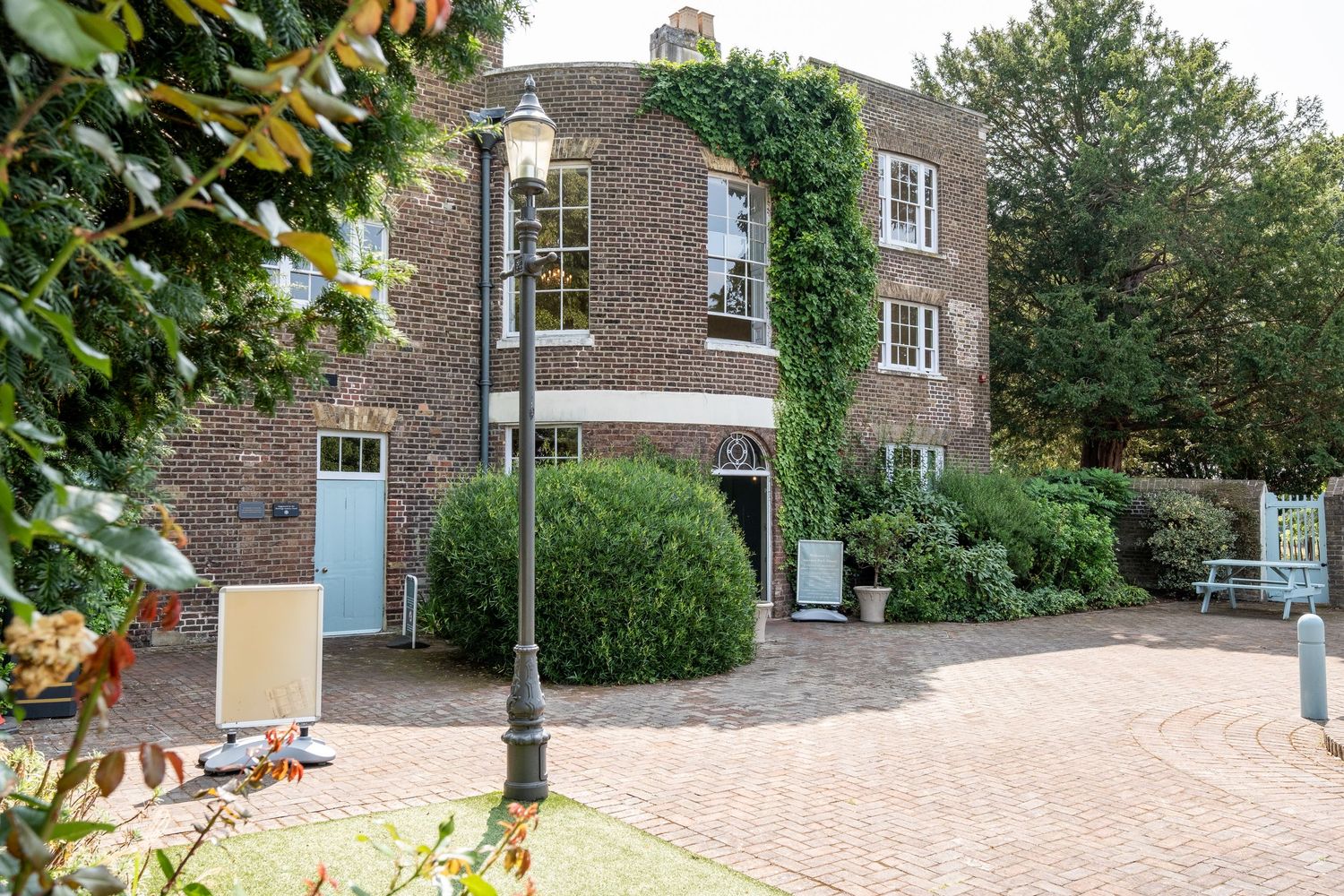 A charming brick building with ivy, blue door, lamp post, and cobblestone courtyard surrounded by lush greenery.