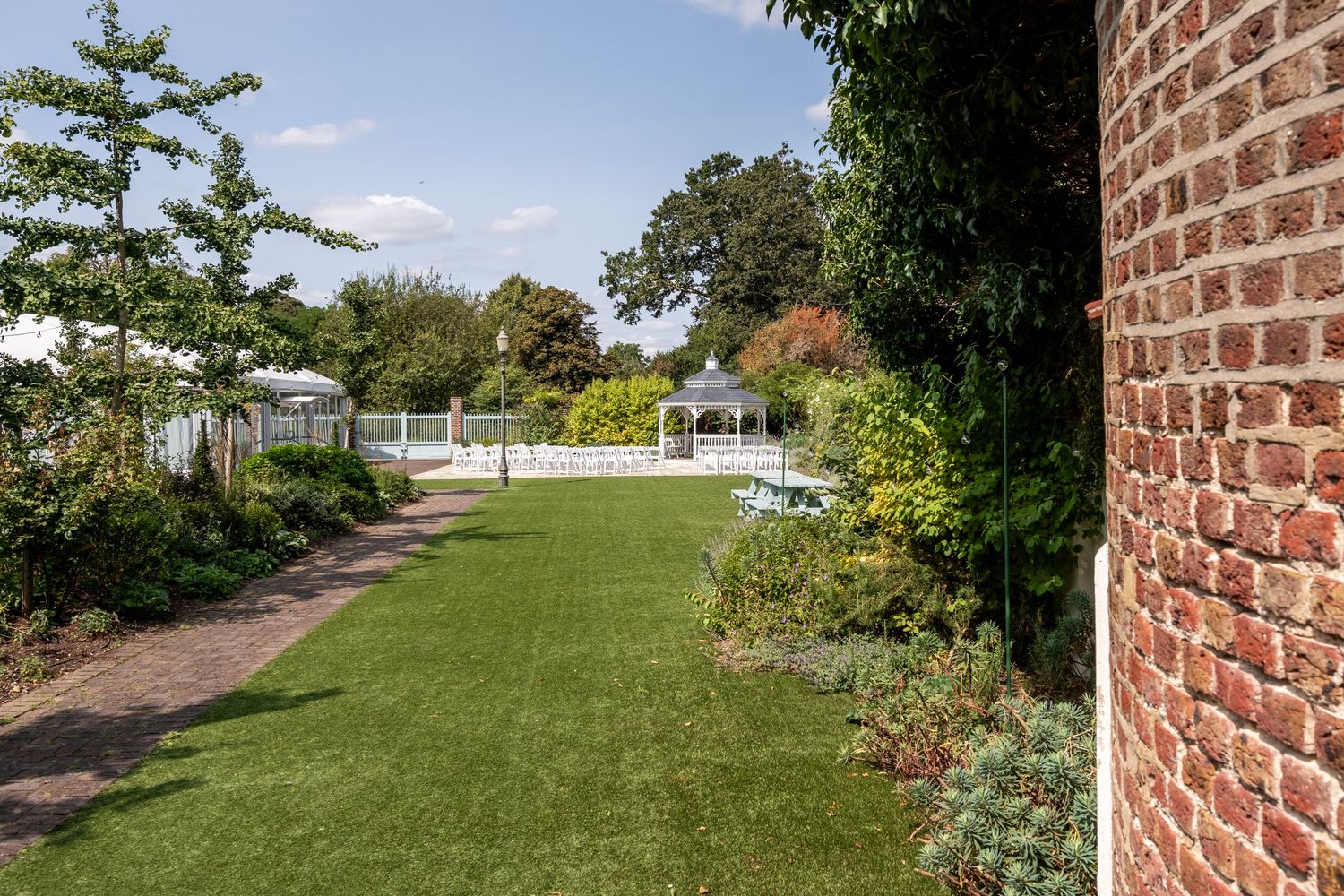Lush green garden with brick wall, manicured lawn, colorful flower borders, white gazebo, and pool area on a sunny day.