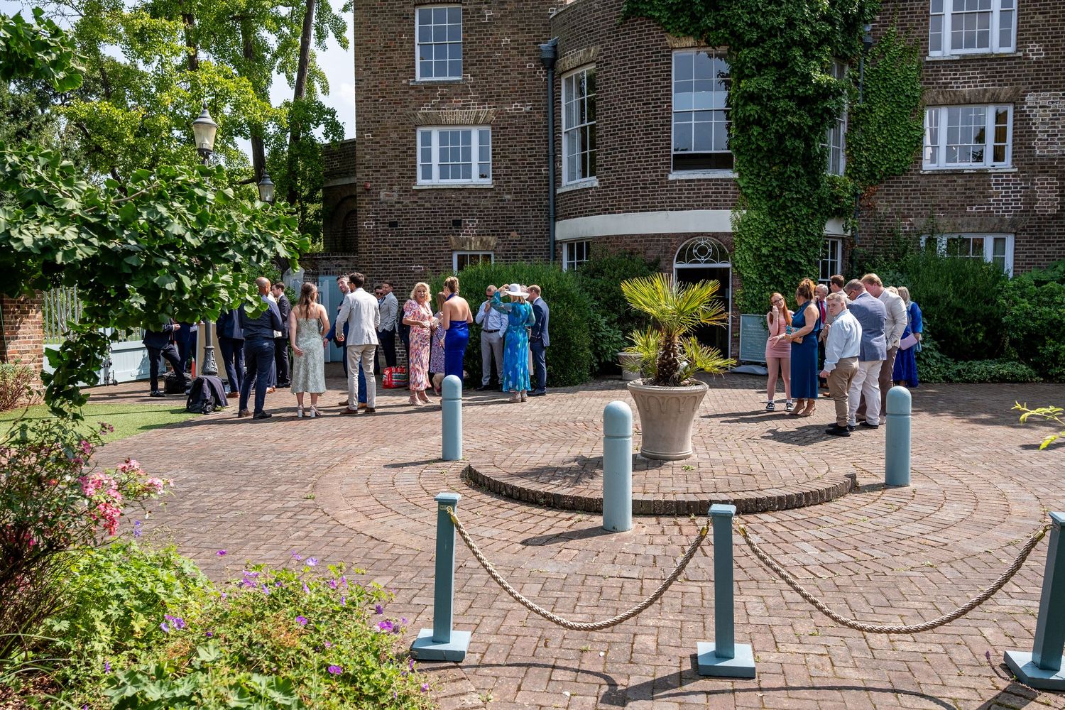 Guests gather on a brick courtyard outside a brick building on a sunny day, surrounded by lush greenery.