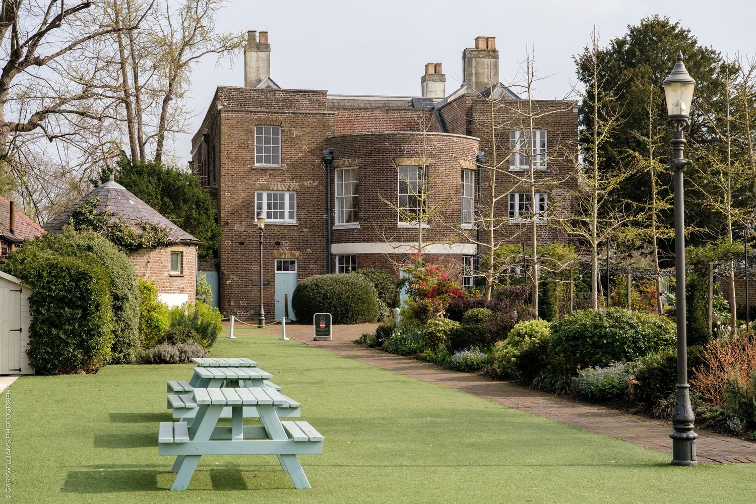 Brick manor house with manicured lawn, picnic benches, ornate lamppost, and lush garden borders on a cloudy day.