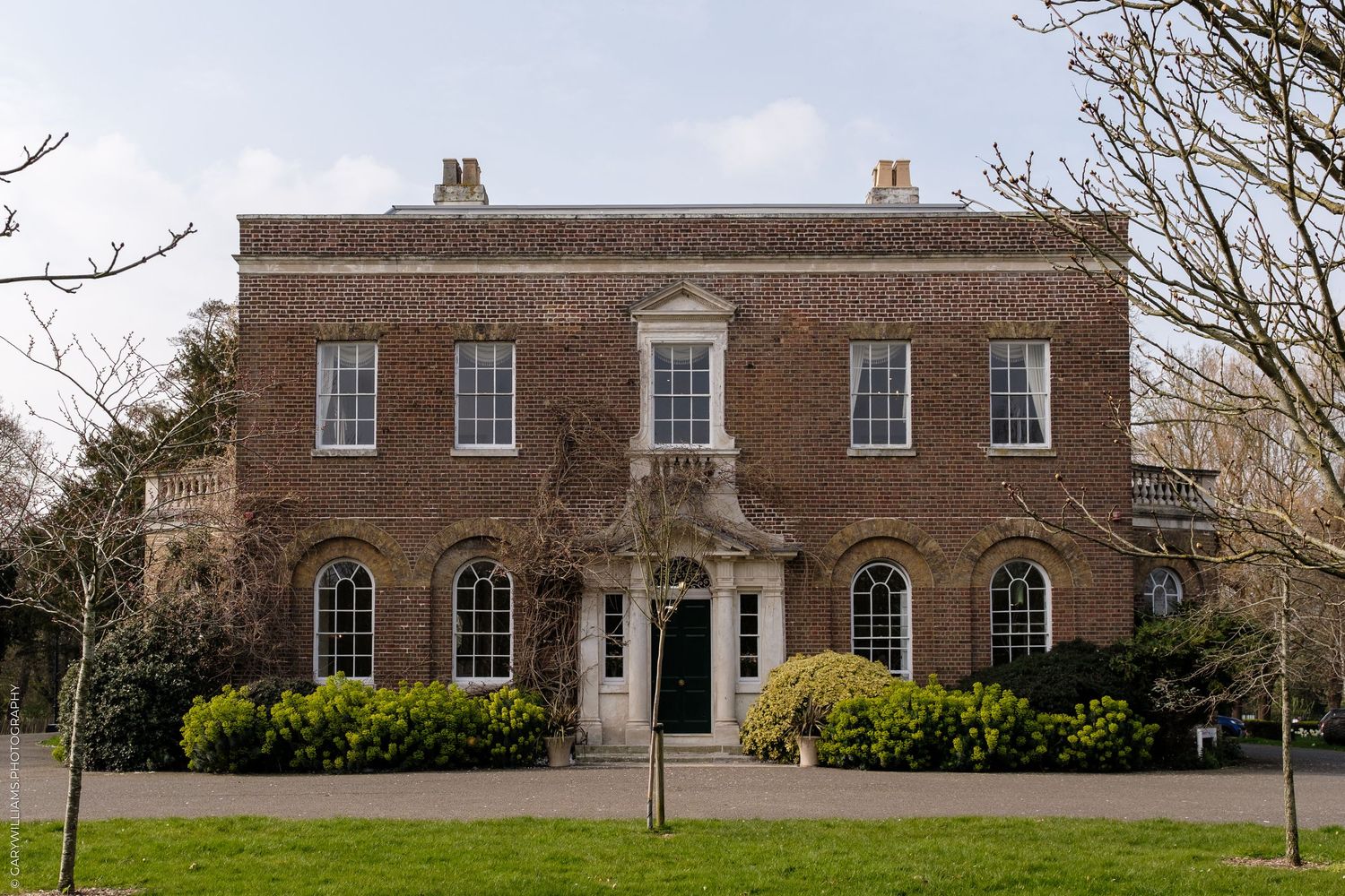 A grand Georgian red-brick manor house with symmetrical windows, ornate entrance, and manicured lawns surrounded by mature trees.