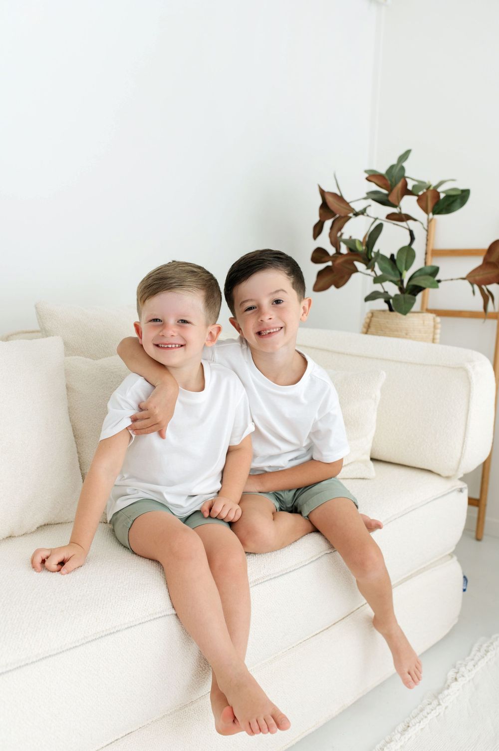 Two smiling boys in matching white tees and green shorts sit together on a cream sofa.
