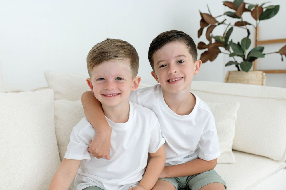 Two smiling boys in white t-shirts sitting together on a cream sofa in a bright living room.