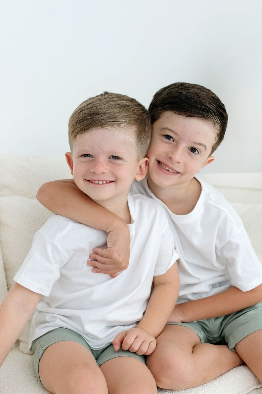 Two smiling boys in white t-shirts and green shorts sit together, with the older boy hugging the younger one.
