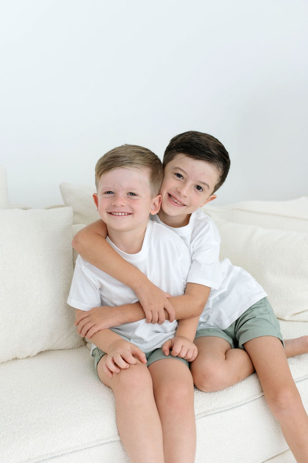 Two smiling boys in white tees hug on a cream sofa.