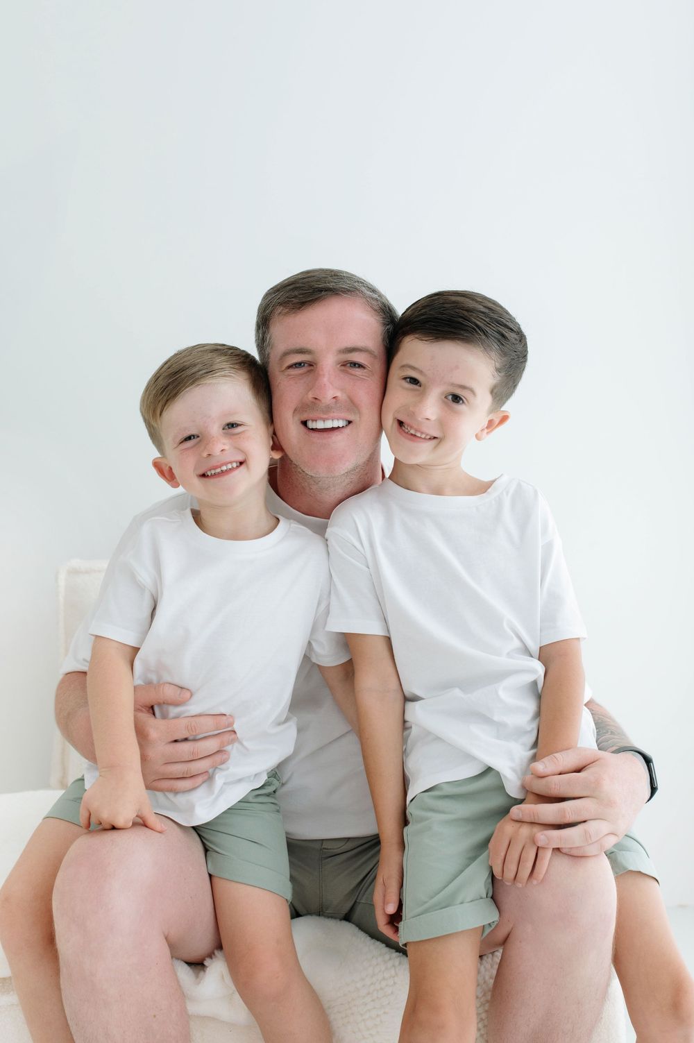 A smiling father sits with his two young sons, all wearing white shirts and green shorts, posing for a portrait.