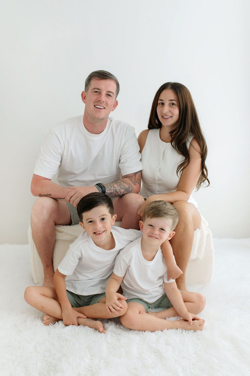 A happy family of four in white outfits poses together on a fluffy white rug against a white background.