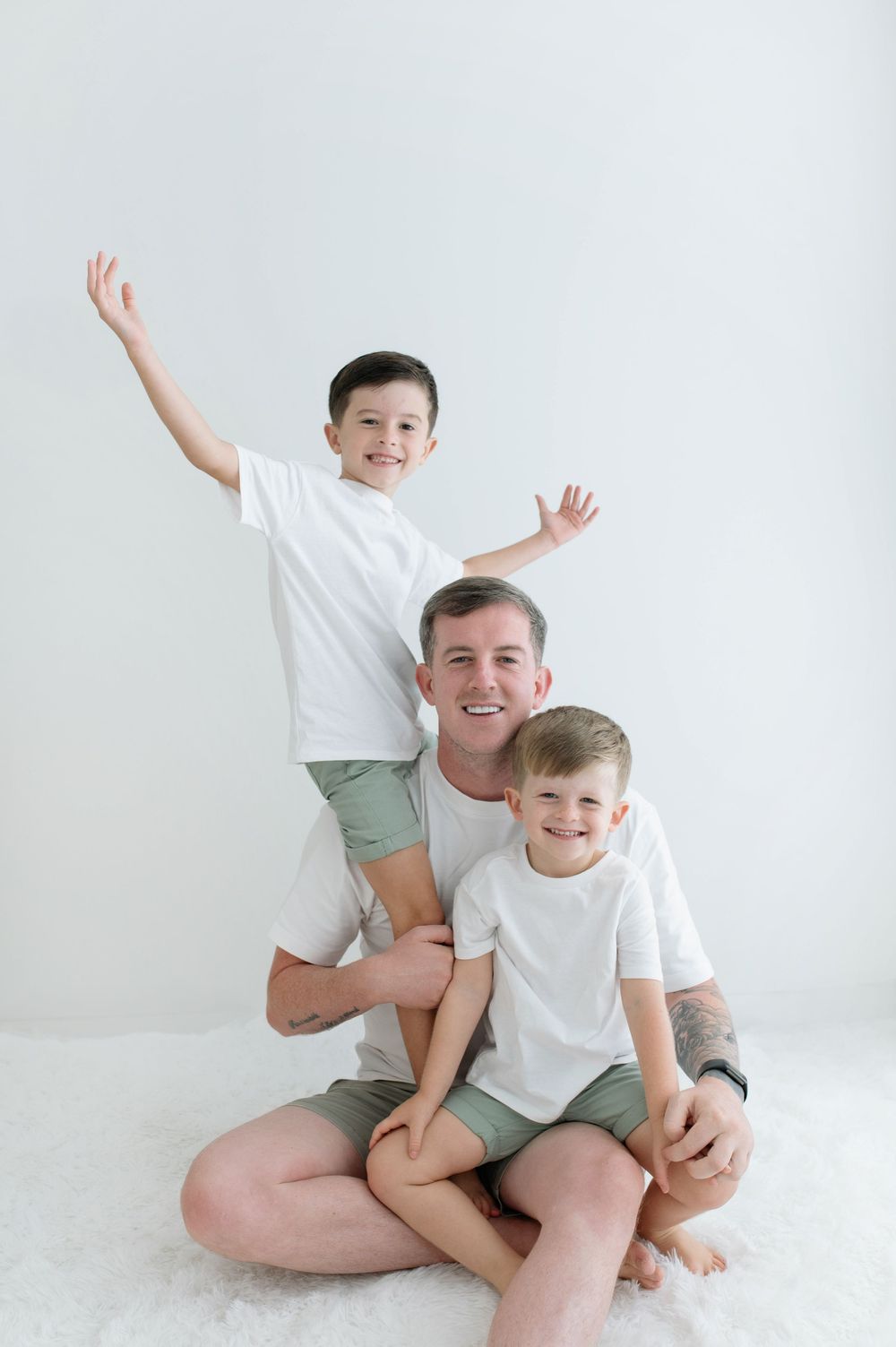 A dad sits cross-legged with two boys in matching white tees and green shorts, all smiling.