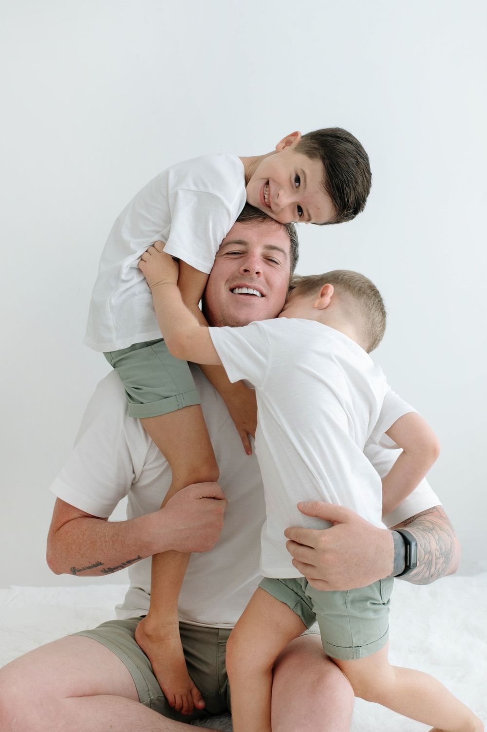 A laughing father hugs his two young sons in matching white tees and green shorts against a white background.
