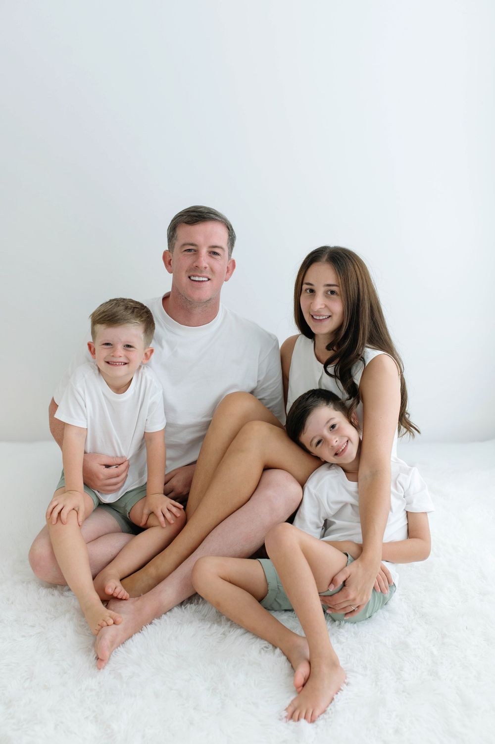 Happy family of four in white outfits sitting together on a white fluffy rug, smiling at the camera.