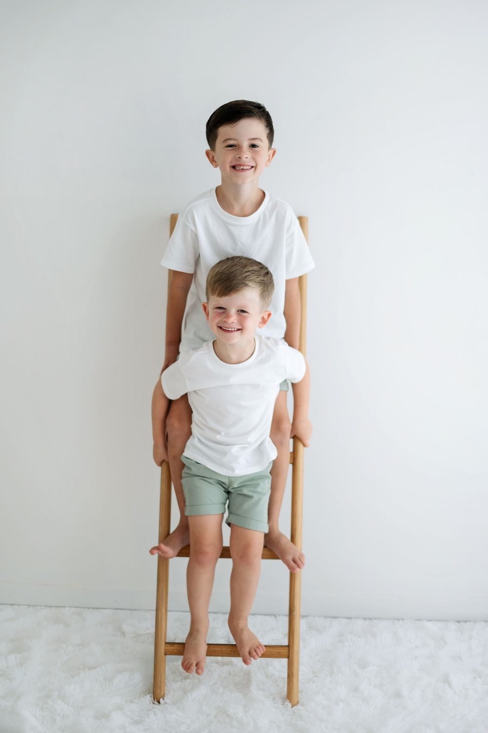 Two smiling brothers in white t-shirts and green shorts pose on a wooden ladder chair against a white wall.