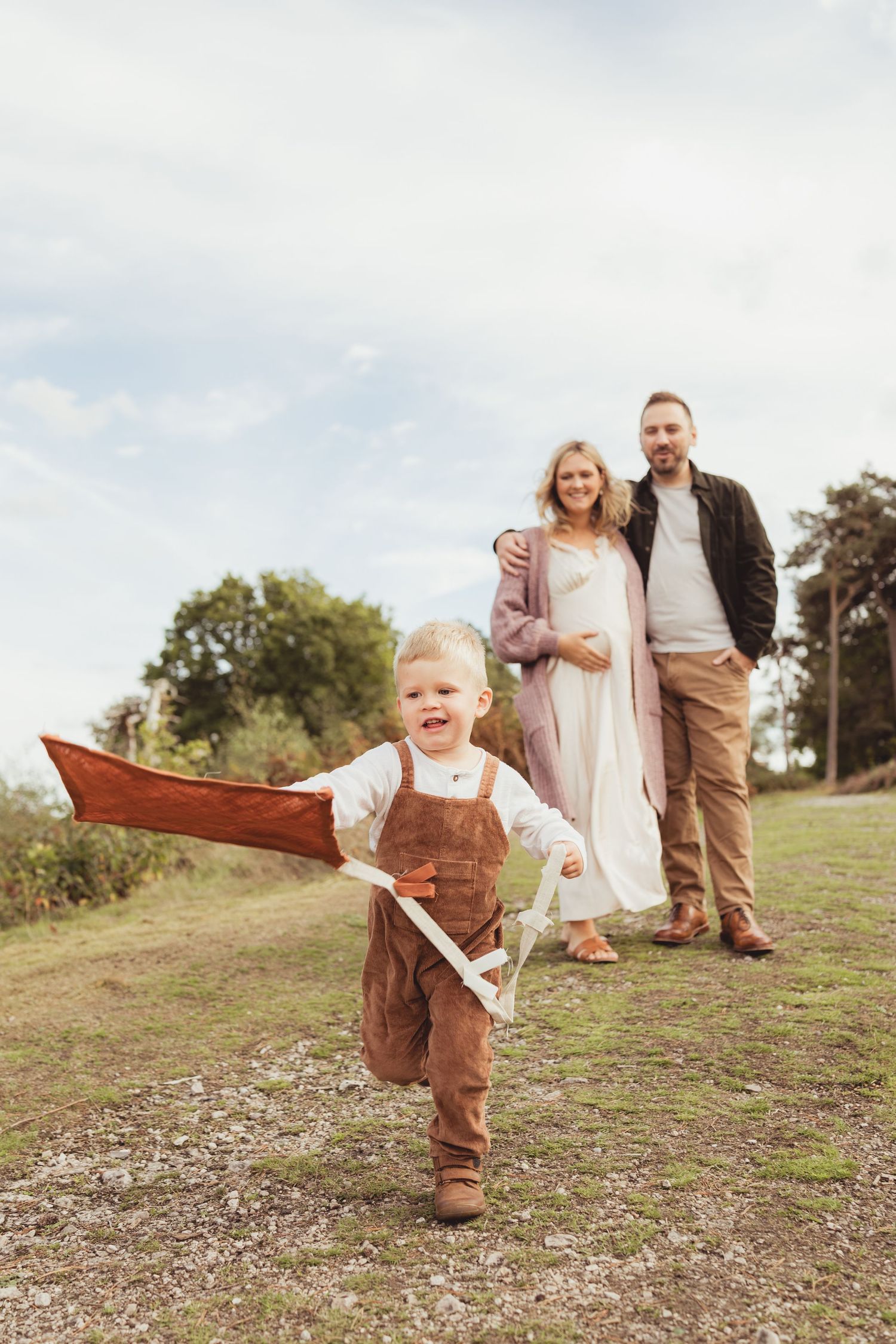A joyful toddler runs with a toy airplane while his parents smile proudly in a scenic outdoor setting.