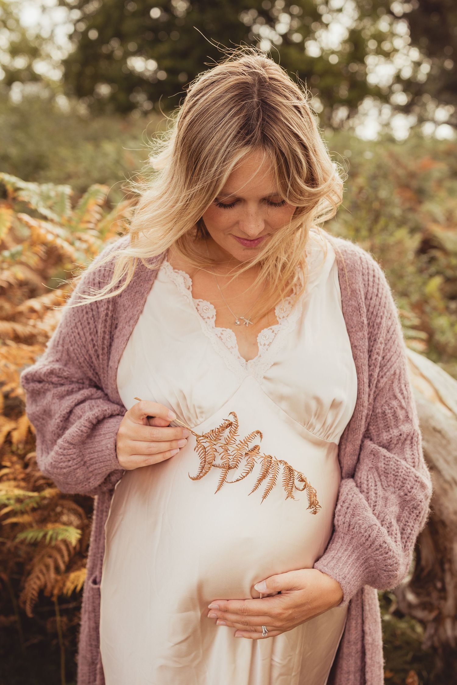 Pregnant woman in white dress and pink cardigan holds golden fern accessory against baby bump outdoors.