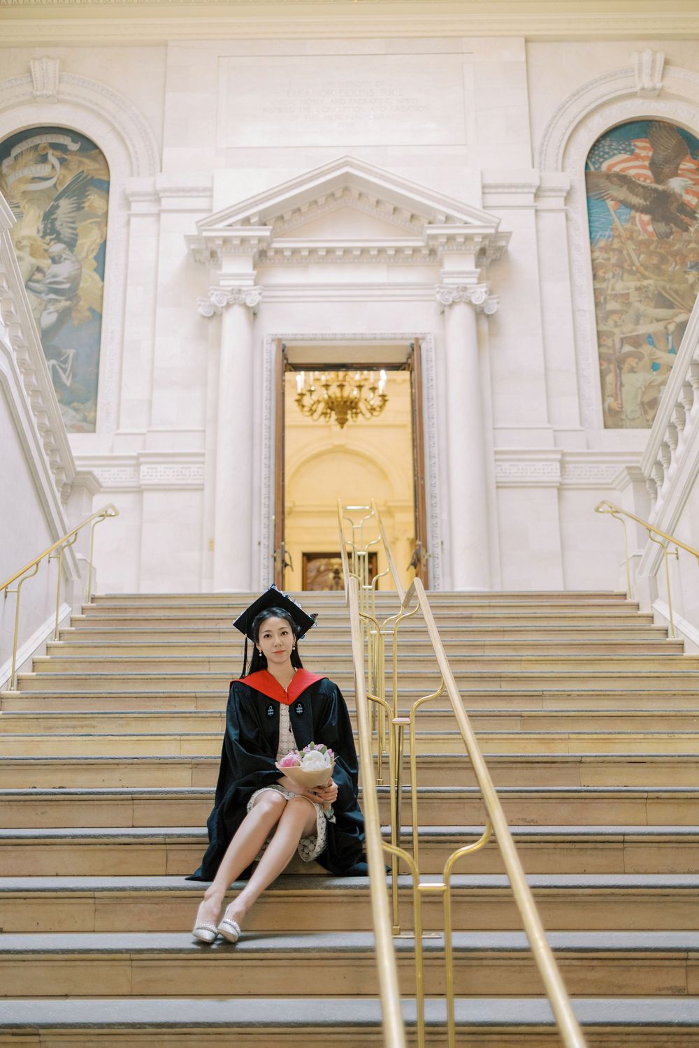 Harvard Graduate in cap and gown sitting on grand marble staircase of Baker Library with ornate murals.