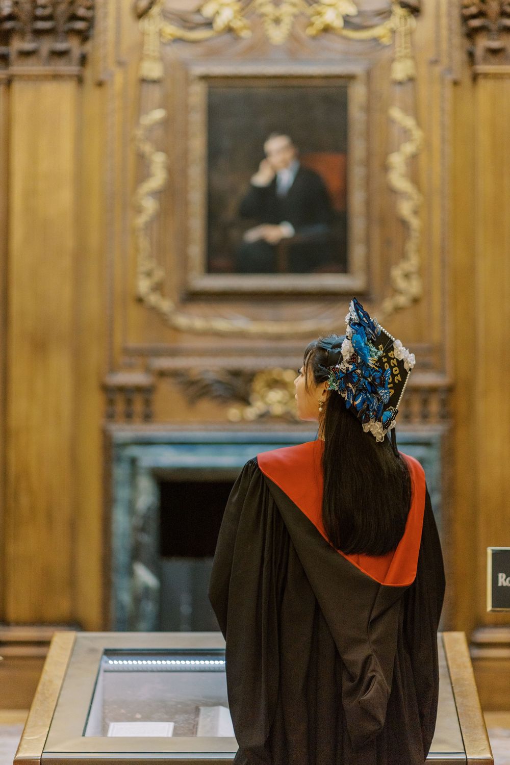 Harvard Graduate in academic gown and decorative cap views a portrait painting in an ornate wood-paneled hall in Baker Library