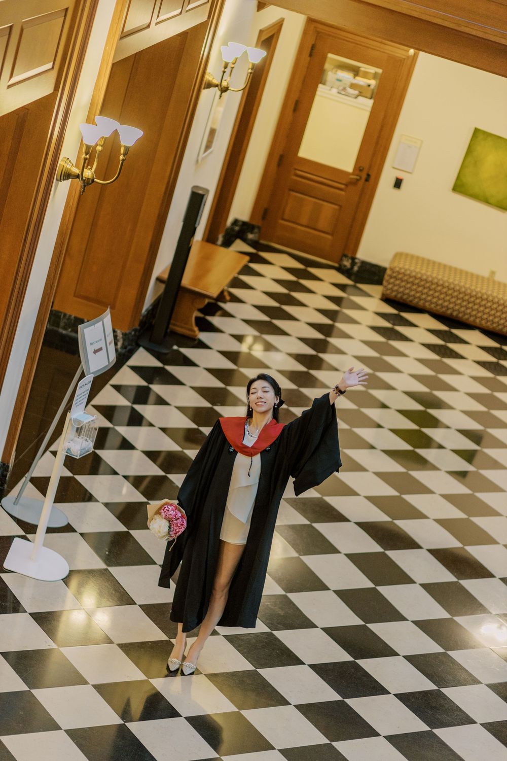 Harvard Graduate in black robe and red sash takes a selfie on a classic black and white checkered floor in Baker Library