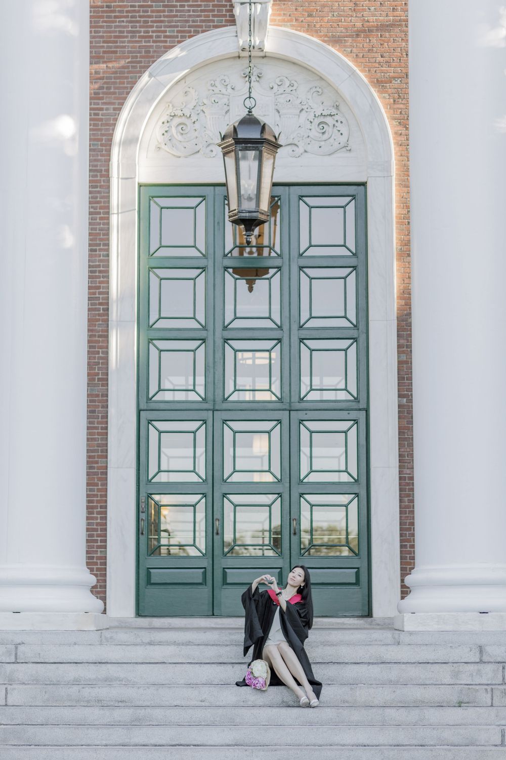 Harvard Graduate in cap and gown sitting on steps of Baker Library before grand green doors with ornate arch and lantern.