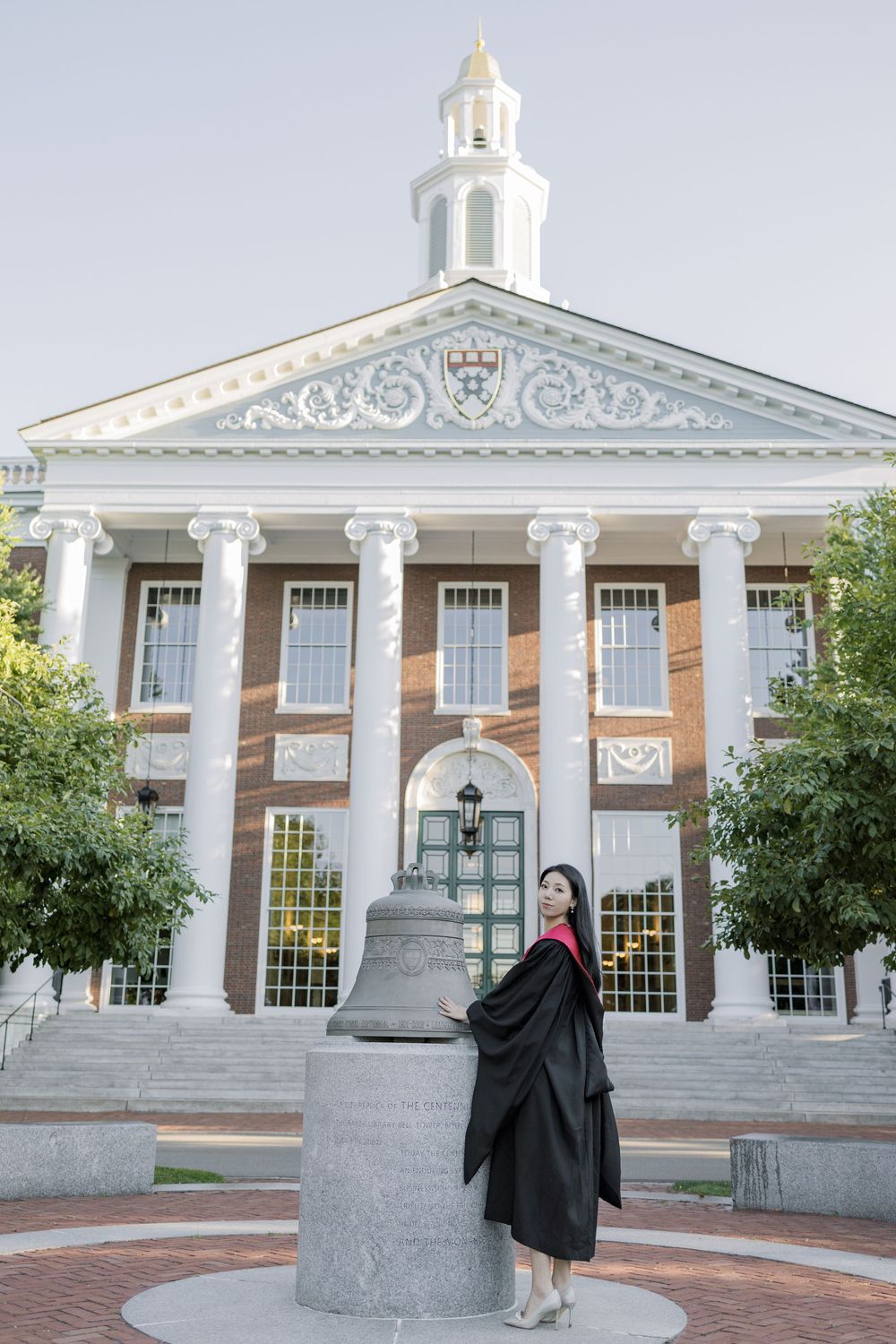 Harvard Graduate in cap and gown posing by a bell in front of a Harvard Business School building.