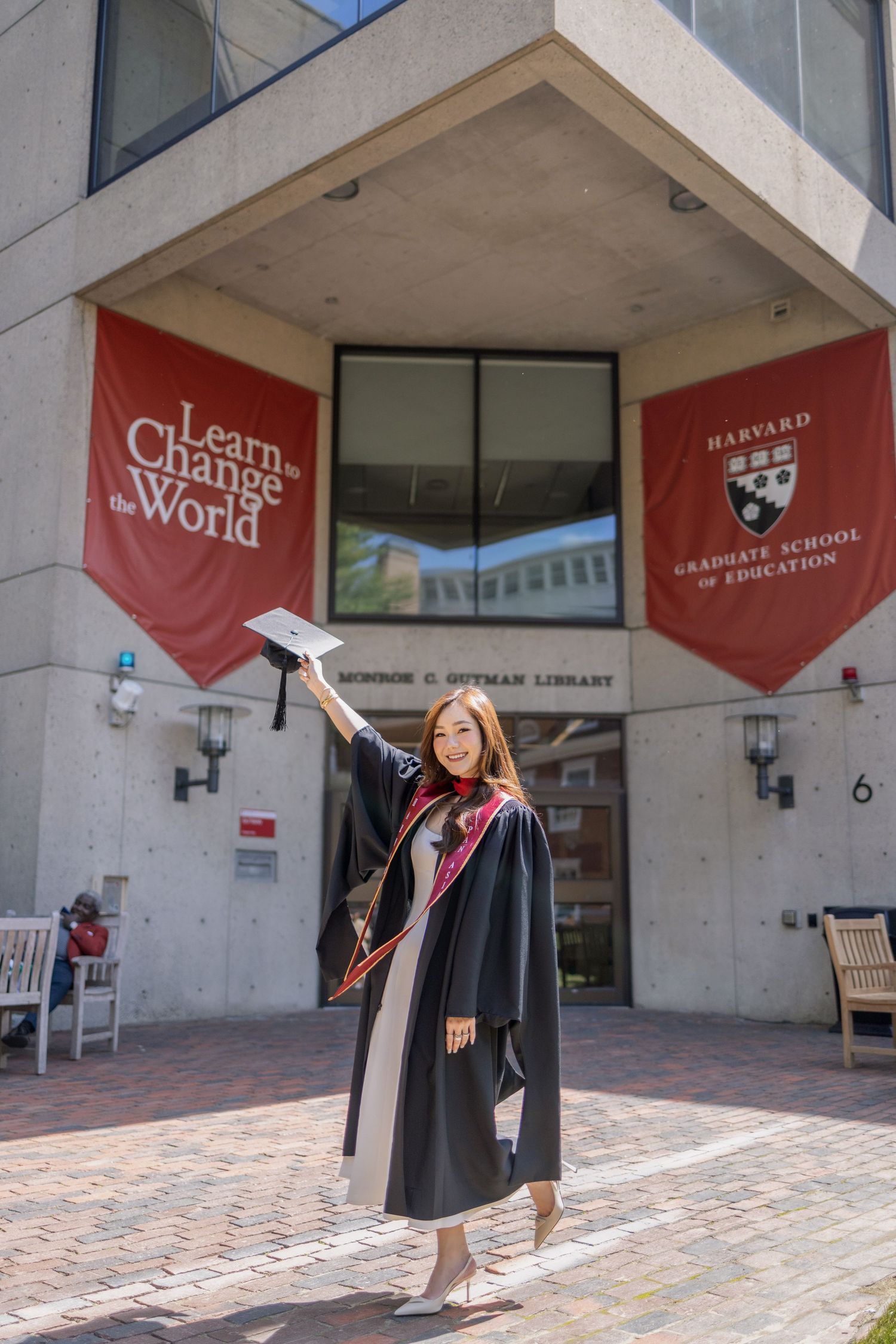 Harvard Graduate School of Education graduate in cap and gown holding mortarboard outside Monroe Gutman Library.