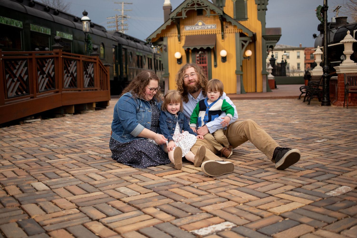 Family of four sitting together on brick pavement outside a vintage train station building.