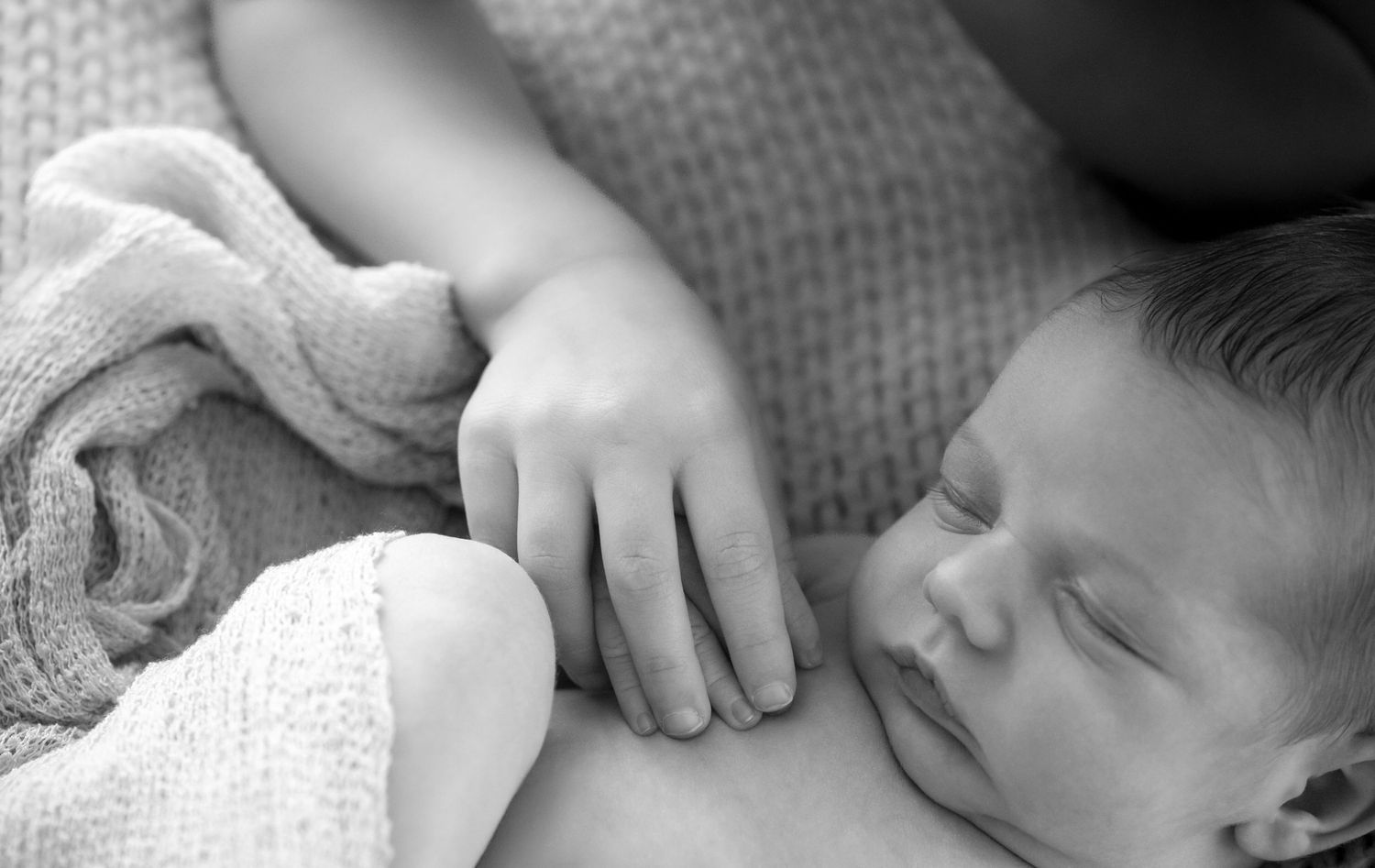 Newborn baby sleeping peacefully while a gentle hand rests on its back, wrapped in a soft pink blanket.