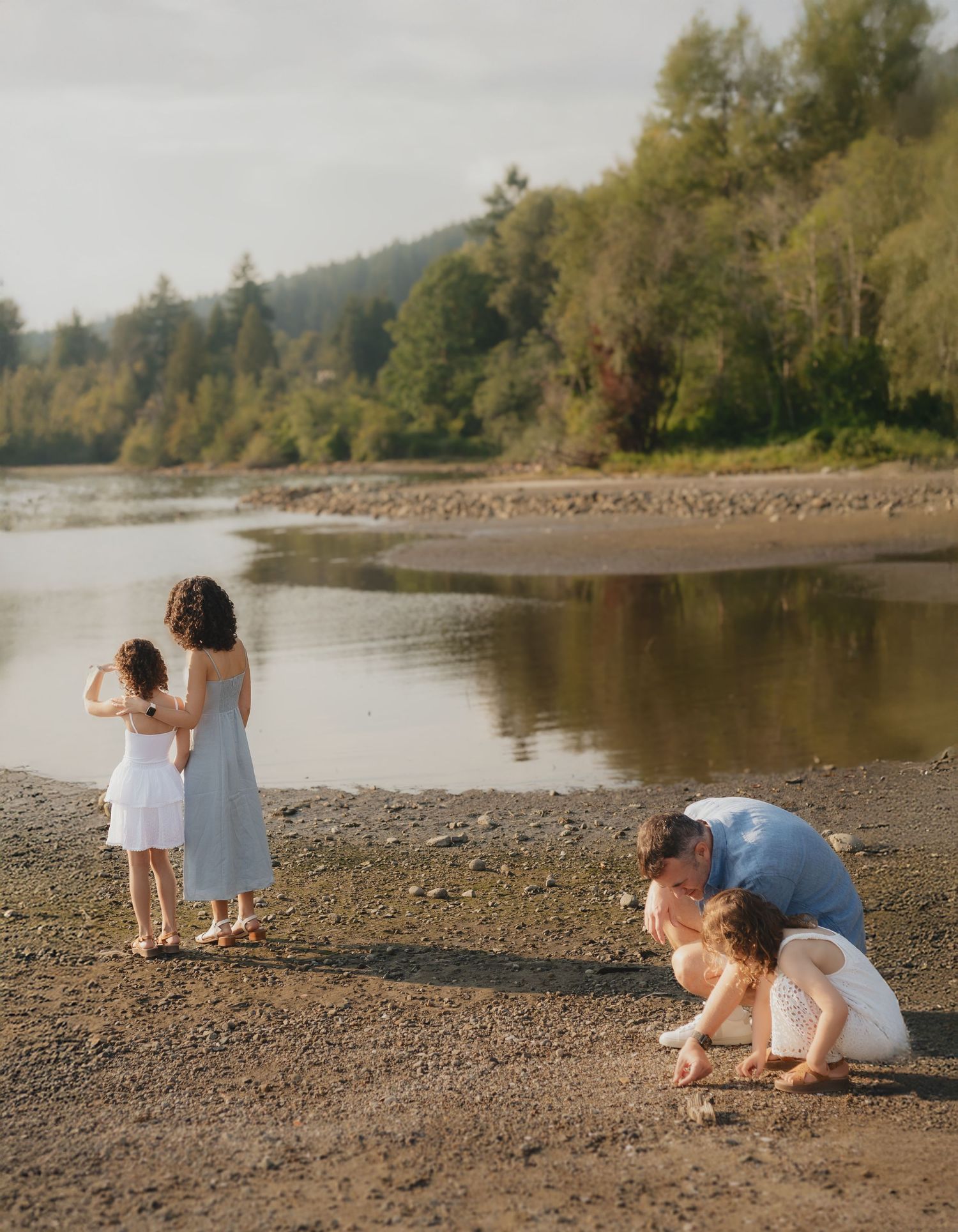 Family enjoying a peaceful day on a rocky riverbank surrounded by lush green trees and calm water.