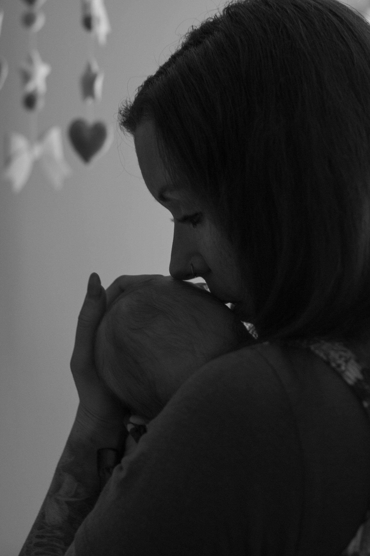 Black and white profile of a mother with tattooed arm gently kissing her newborn baby near a hanging mobile.