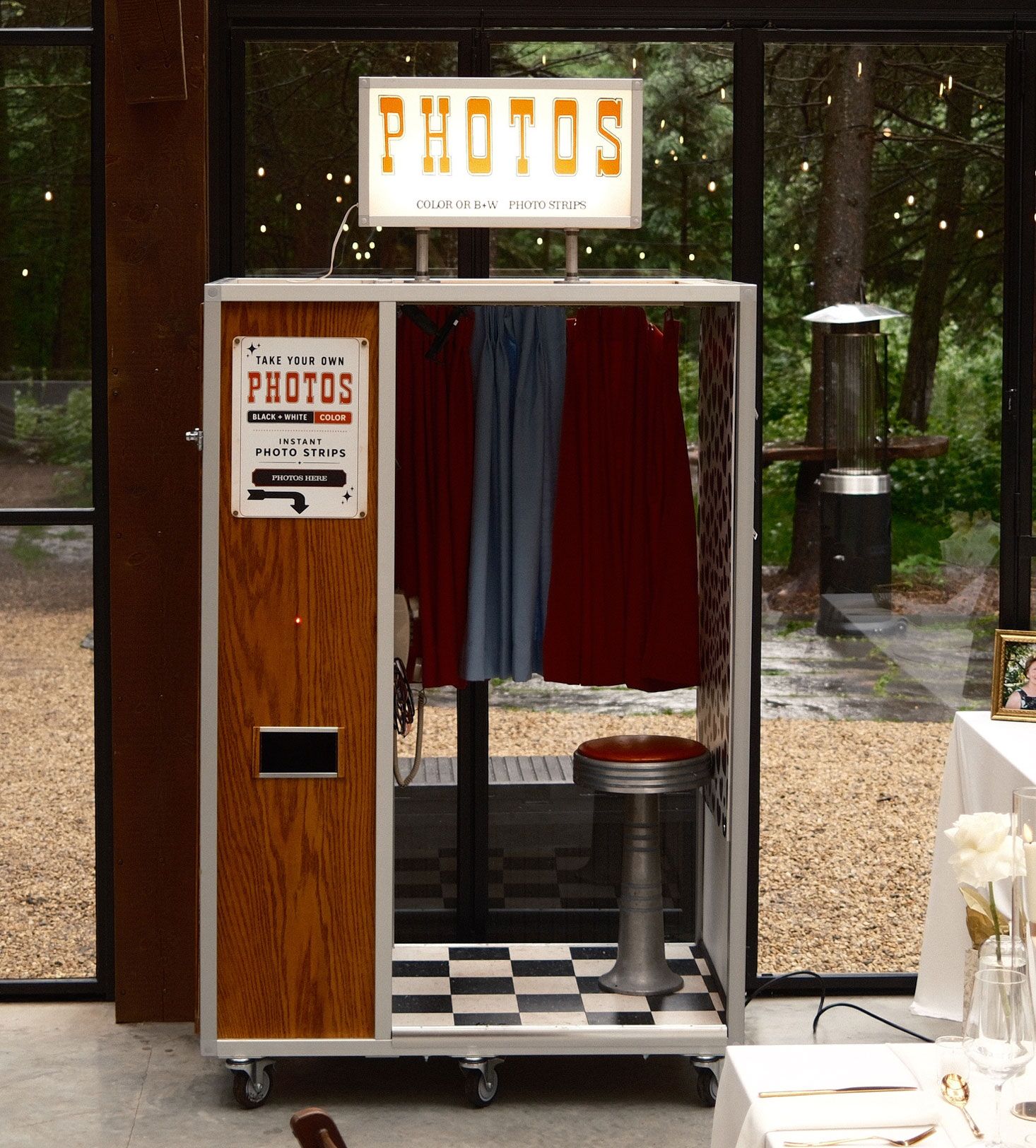 Retro photo booth with checkered floor, heart wallpaper, orange curtain, stool, and color or B&W photo strips sign.