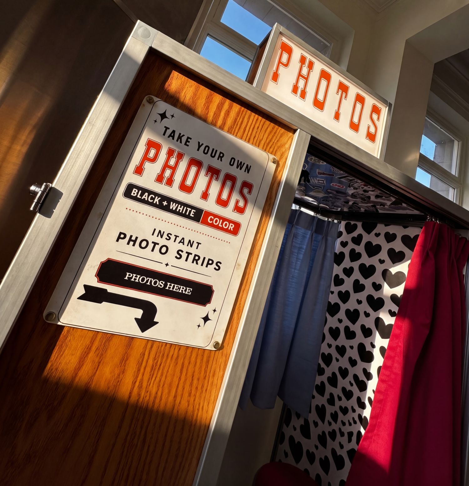 Retro photo booth with checkered floor, heart wallpaper, orange curtain, stool, and color or B&W photo strips sign.