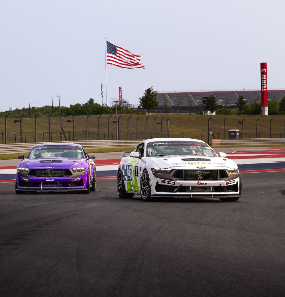 Two race cars, purple and white Ford Mustangs, on a track with an American flag waving in the background.