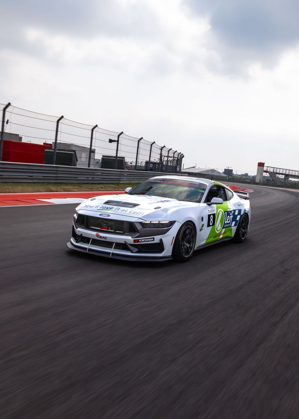 White Ford Mustang GT4 race car with green accents speeding on a professional racing circuit track.