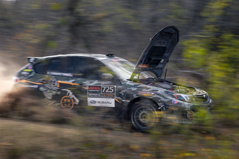 Subaru rally car number 725 speeding through a dirt forest stage with hood open and dust flying.