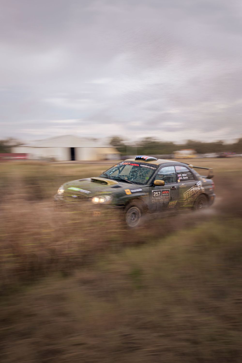 Rally car number 257 speeds through a muddy off-road track during a motorsport event on an overcast day.