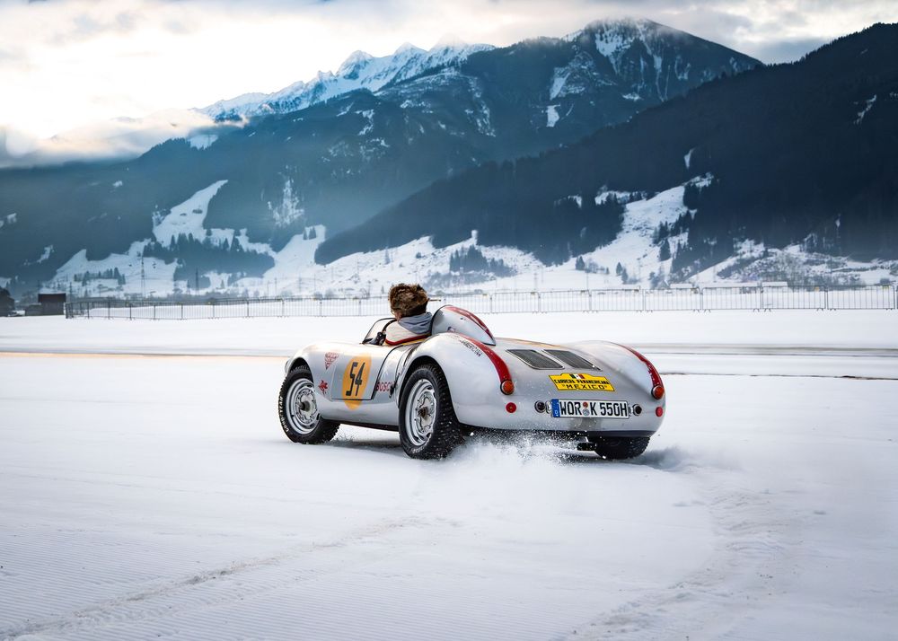 Vintage racing car driving on snowy track with snow-capped Alpine mountains in background during winter.