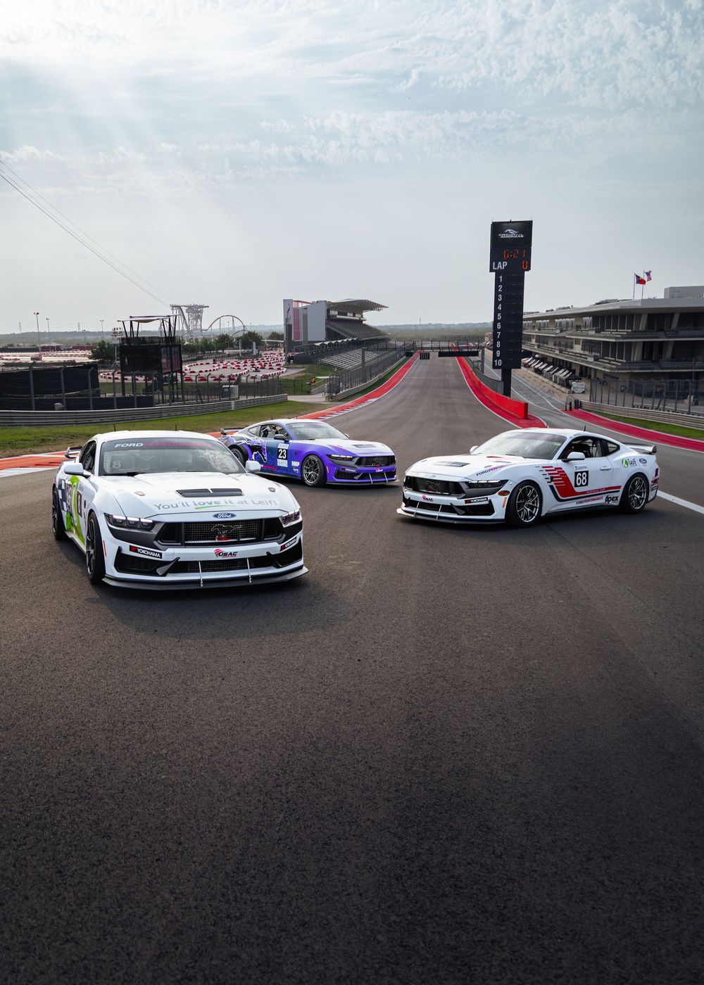 Three Ford Mustang race cars posed on a motorsport track circuit under dramatic skies at Circuit of the Americas.