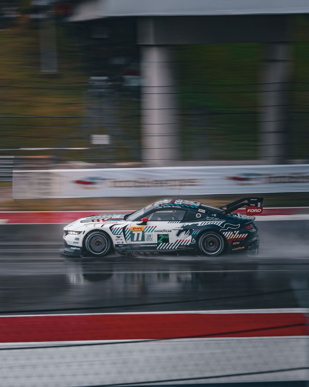 A Ford Mustang GT4 race car speeds through a wet track in rainy conditions during a motorsport event.