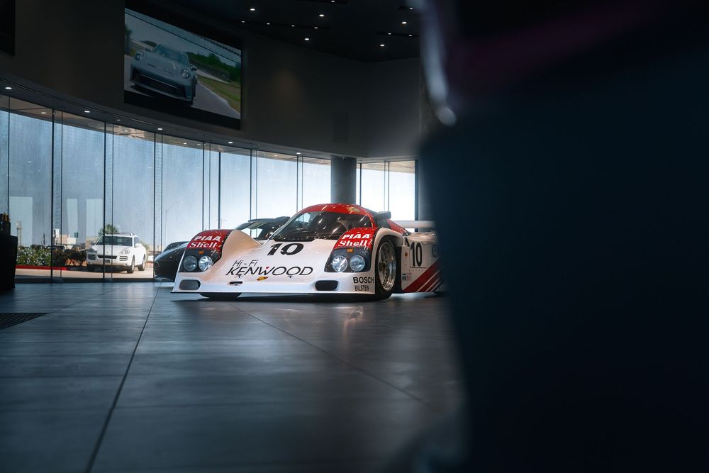 Porsche 962 race car with Kenwood livery displayed in a modern museum showroom.