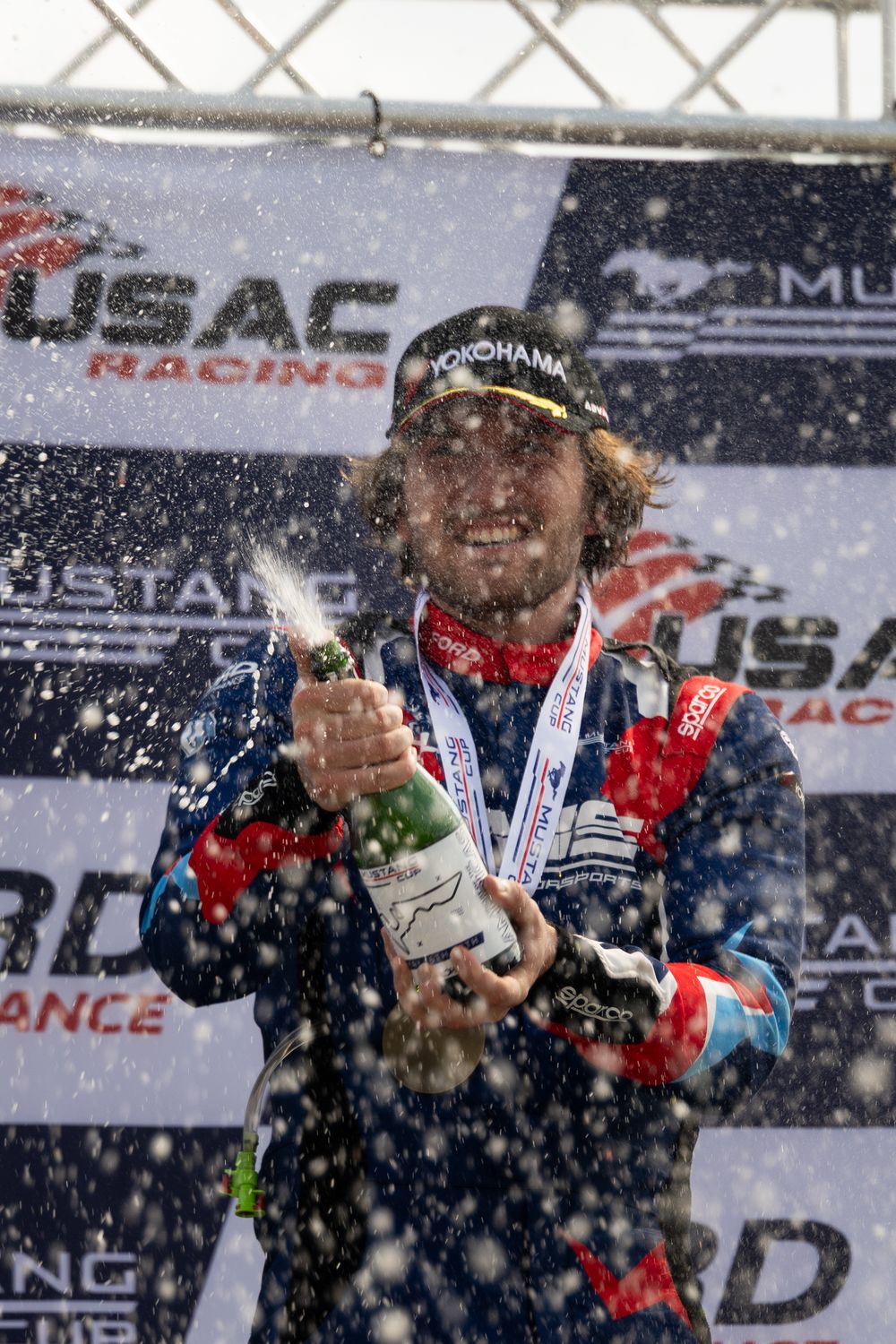 USAC Racing winner celebrates on podium spraying champagne wearing Yokohama cap and race suit with medal.